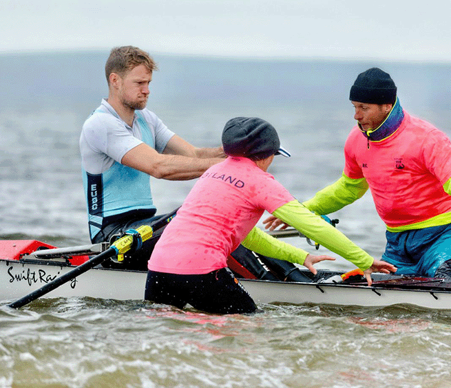 Coastal sculler launching with help of boat handlers, all illustrating what to wear for coastal sculling